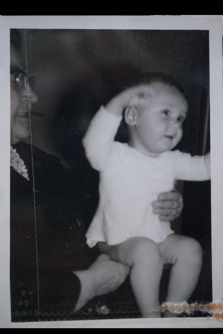 Schwarz/Weiß-Foto. Man sieht ein Baby im weißem Hemdchen sitzend neben seiner Oma.